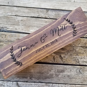 May include: A rectangular wooden sign with the names "Jenna & Matt" and the date "July 21, 2018" engraved in a script font. The sign is stained a dark brown and features leaf designs on each end.
