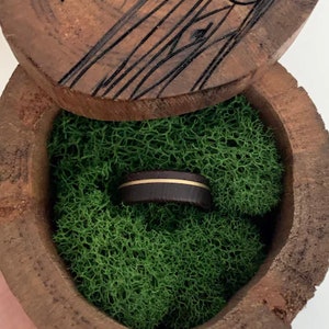 K&ouml;nnte beinhalten: Eine Holzkiste f&uuml;r Ringe mit gr&uuml;ner Moos-Auskleidung. Ein dunkelbrauner Ring mit einem d&uuml;nnen wei&szlig;en Streifen befindet sich in der Kiste.