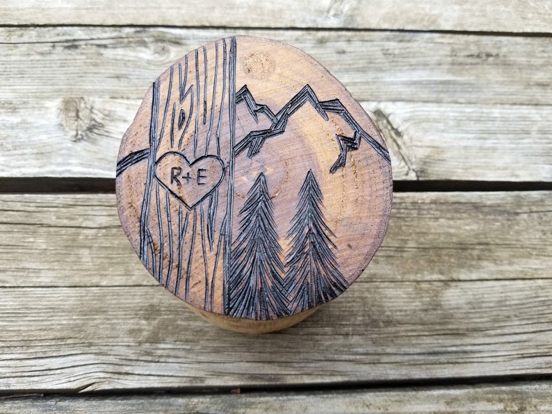 K&ouml;nnte beinhalten: Eine runde Holzscheibe mit einem geschnitzten Design aus zwei B&auml;umen, Bergen und einem Herzen mit den Initialen "R+E" darin.