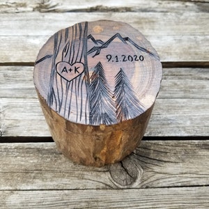 K&ouml;nnte beinhalten: Eine Holzkiste f&uuml;r Ringe mit einem geschnitzten Design aus B&auml;umen, Bergen und einem Herzen mit den Initialen "A+K" und dem Datum "9.1.2020".