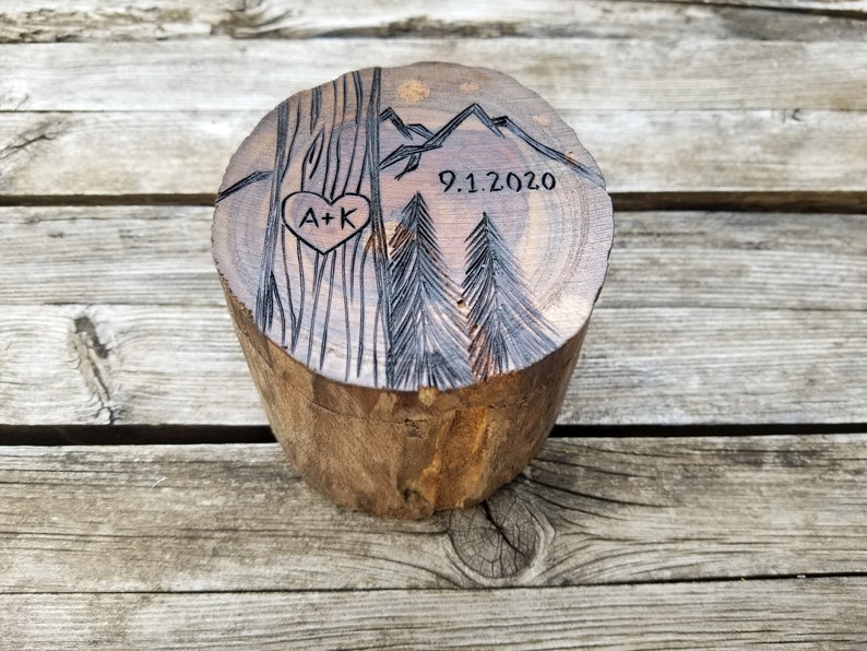 K&ouml;nnte beinhalten: Eine Holzkiste f&uuml;r Ringe mit einem geschnitzten Design aus B&auml;umen, Bergen und einem Herzen mit den Initialen "A+K" und dem Datum "9.1.2020".