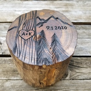 K&ouml;nnte beinhalten: Eine Holzkiste f&uuml;r Ringe mit einem geschnitzten Design aus Bergen, B&auml;umen und einem Herzen mit den Initialen "A+K" darin. Das Datum "9.1.2020" ist ebenfalls in das Holz geschnitzt.