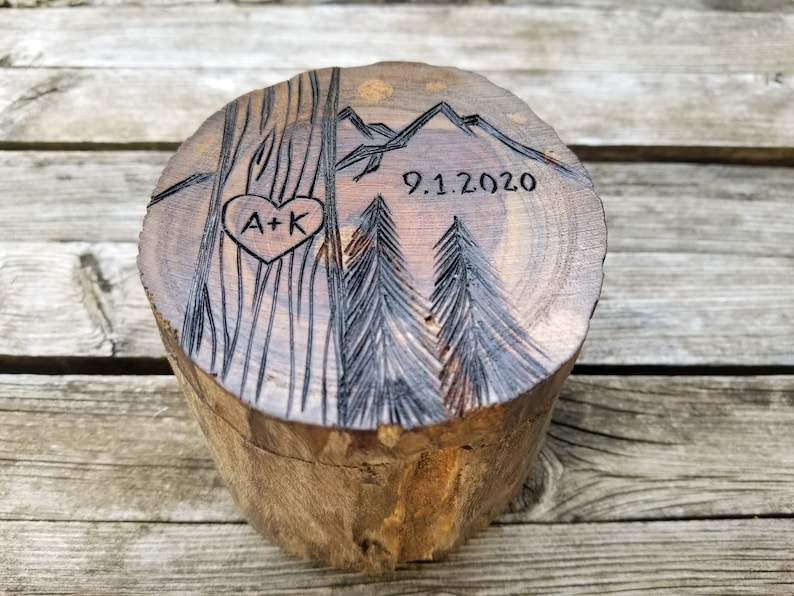 K&ouml;nnte beinhalten: Eine Holzkiste f&uuml;r Ringe mit einem geschnitzten Design aus Bergen, B&auml;umen und einem Herzen mit den Initialen "A+K" darin. Das Datum "9.1.2020" ist ebenfalls in das Holz geschnitzt.