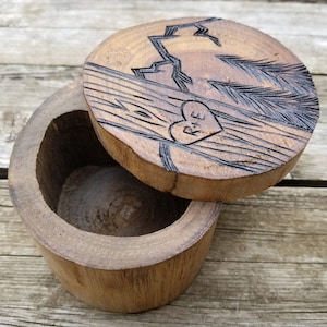 K&ouml;nnte beinhalten: Eine Holzkiste f&uuml;r Ringe mit einem geschnitzten Design aus einer Bergkette und Kiefern. Der Deckel hat ein Herz mit den Initialen "R+E" darin geschnitzt.