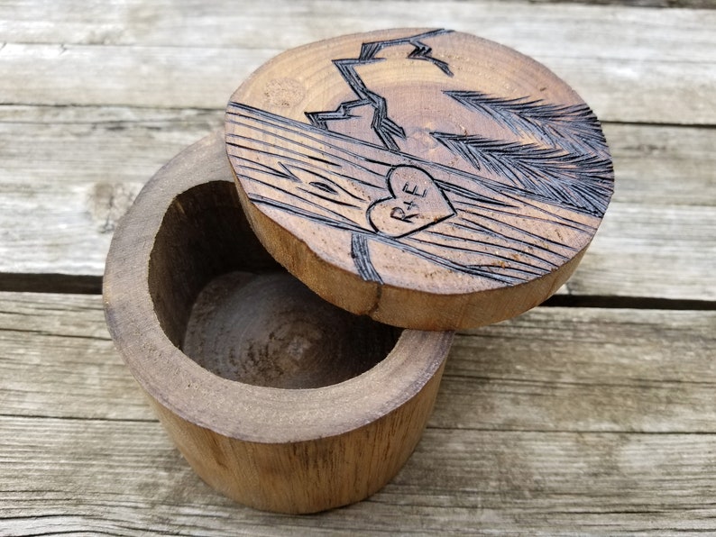 K&ouml;nnte beinhalten: Eine Holzkiste f&uuml;r Ringe mit einem geschnitzten Design aus einer Bergkette und Kiefern. Der Deckel hat ein Herz mit den Initialen "R+E" darin geschnitzt.