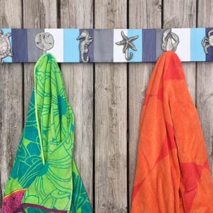 May include: A wooden towel rack with five hooks shaped like sea creatures. The hooks are painted silver and the rack is painted in alternating stripes of blue, white, and gray. Two towels are hanging on the hooks, one green and one orange.
