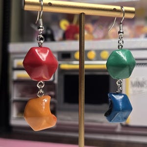 Puede incluir: Un par de pendientes coloridos exhibidos en un soporte dorado. Cada pendiente presenta una forma geométrica roja y naranja, y una forma geométrica verde y azul. Los pendientes están suspendidos de ganchos plateados.