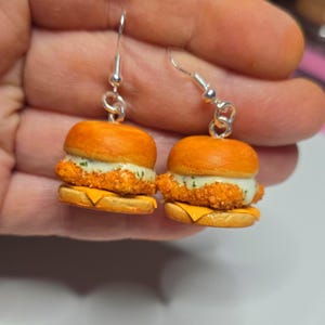 Puede incluir: Un par de pendientes novedosos con sándwiches de pollo en miniatura. Cada pendiente tiene un gancho plateado y una pequeña hamburguesa con pan dorado, pollo crujiente, queso y una salsa cremosa.