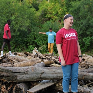 May include: Three people standing in a wooded area near a stream. The person in the foreground is wearing a red t-shirt with the text "HUGS SEE BACK FOR DETAIL" printed on it. They are standing on a pile of logs and rocks.