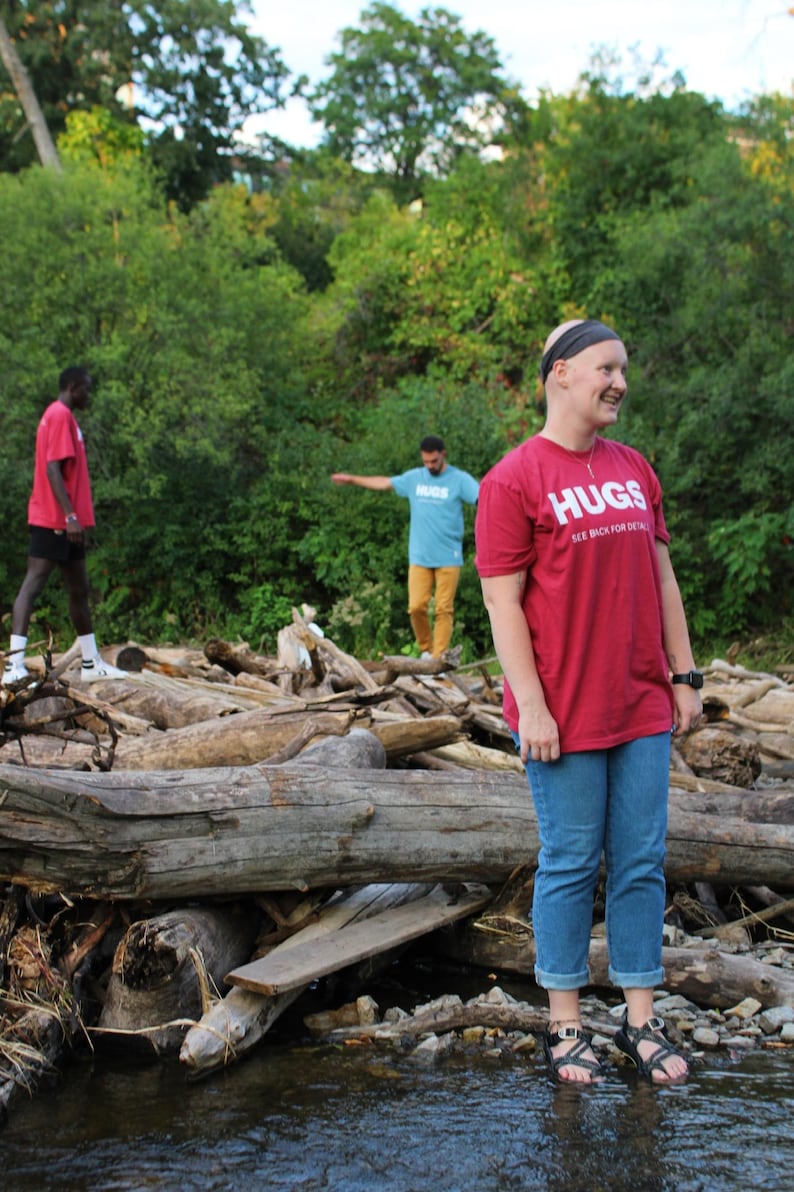 May include: Three people standing in a wooded area near a stream. The person in the foreground is wearing a red t-shirt with the text "HUGS SEE BACK FOR DETAIL" printed on it. They are standing on a pile of logs and rocks.