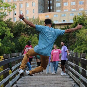 May include: A group of people standing on a wooden bridge. One person is jumping in the air with their arms outstretched. They are wearing a blue t-shirt with the text "Spread the Love" and brown pants. The other people are wearing various colored shirts and pants. The bridge is surrounded by a wooden railing.