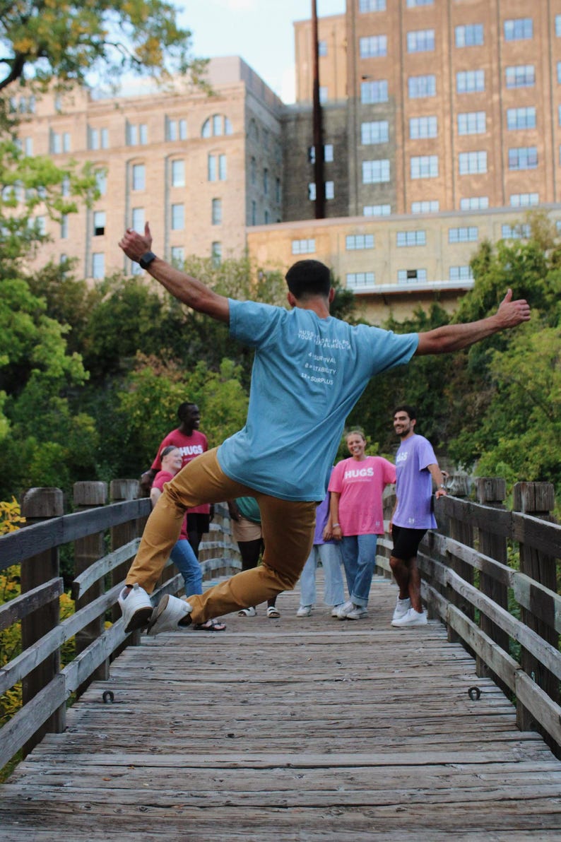 May include: A group of people standing on a wooden bridge. One person is jumping in the air with their arms outstretched. They are wearing a blue t-shirt with the text "Spread the Love" and brown pants. The other people are wearing various colored shirts and pants. The bridge is surrounded by a wooden railing.