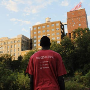 May include: A person wearing a red t-shirt with the text "Hugs per day helps your mental health 4=survival 8=stability 12=surplus" in white. The person is standing in front of a brick building with a sign that says "Miller's", and there are trees in the background.