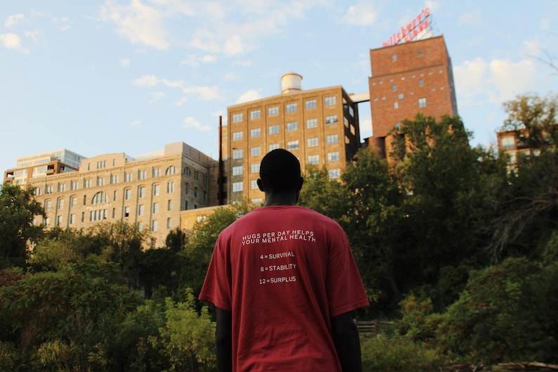 May include: A person wearing a red t-shirt with the text "Hugs per day helps your mental health 4=survival 8=stability 12=surplus" in white. The person is standing in front of a brick building with a sign that says "Miller's", and there are trees in the background.