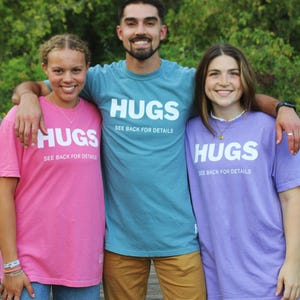 May include: Three people wearing matching t-shirts with the word "HUGS" and the text "See back for details". The shirts are pink, teal, and purple.
