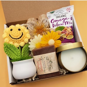 May include: A gift box filled with a crocheted sunflower, a candle, a bar of soap, and a bag of organic omega-3 mix. The box is lined with brown paper and has a white background.