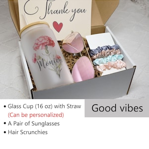 May include: A white gift box containing a personalised glass cup with a straw, a pair of pink sunglasses, and hair scrunchies in various colours. The box is filled with brown packing paper and the text "Thank you" and "Monica" are visible on the box. The text "Good vibes" is visible below the box.