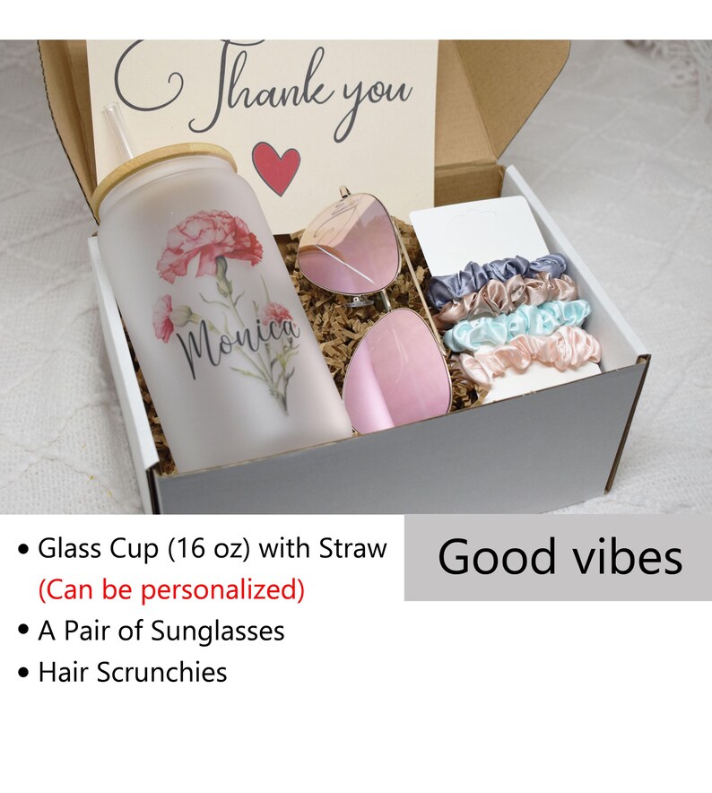 May include: A white gift box containing a personalised glass cup with a straw, a pair of pink sunglasses, and hair scrunchies in various colours. The box is filled with brown packing paper and the text "Thank you" and "Monica" are visible on the box. The text "Good vibes" is visible below the box.