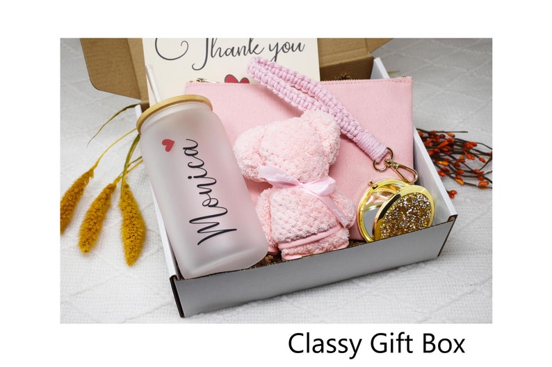 May include: A white gift box filled with pink and gold items. The box contains a pink fuzzy teddy bear, a pink pouch, a clear glass water bottle with a bamboo lid and the name "Monica" written on it, and a gold compact mirror with rhinestones.