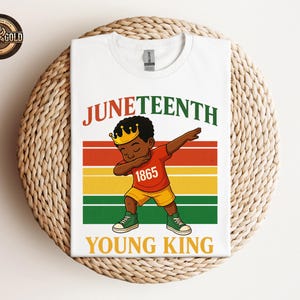 Puede incluir: Camiseta blanca con una ilustraci&oacute;n de dibujos animados de un ni&ntilde;o negro joven haciendo dab, con una corona y el n&uacute;mero 1865. La camiseta tiene las palabras "Juneteenth" y "Young King" en un dise&ntilde;o colorido.