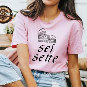 May include: A light pink t-shirt featuring a black line drawing of the Colosseum and the words "sei sette" in black script. The shirt has short sleeves and is worn by a person. A "Rose Gold" logo is in the upper left corner.