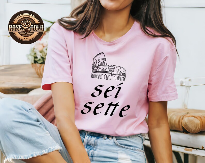 May include: A light pink t-shirt featuring a black line drawing of the Colosseum and the words "sei sette" in black script. The shirt has short sleeves and is worn by a person. A "Rose Gold" logo is in the upper left corner.