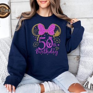 May include: Navy blue sweatshirt with a glittery pink bow and "50th Birthday" text. The design includes gold accents, fireworks, and a diamond. A crown and castle are also featured in the design.