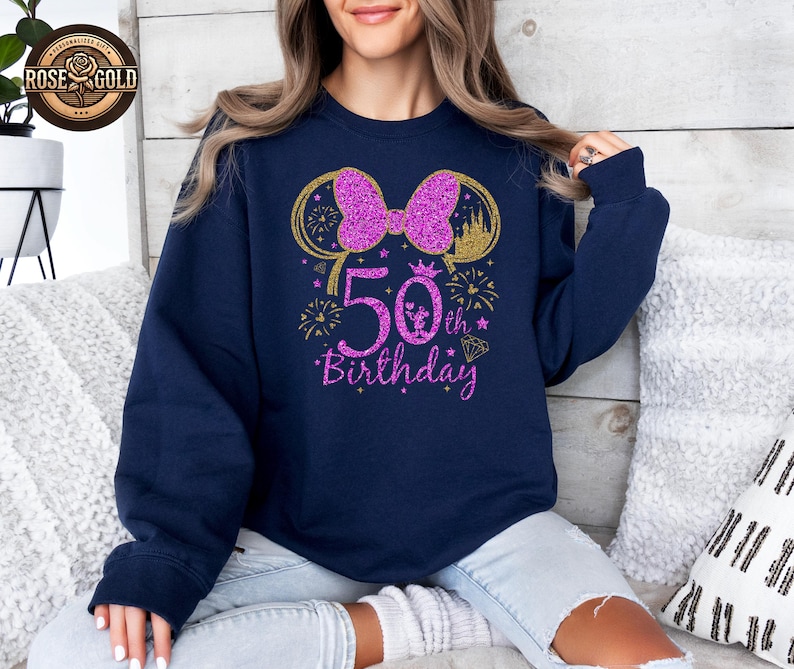 May include: Navy blue sweatshirt with a glittery pink bow and "50th Birthday" text. The design includes gold accents, fireworks, and a diamond. A crown and castle are also featured in the design.