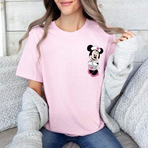 Puede incluir: Una camiseta rosa claro con un dise&ntilde;o de Minnie Mouse. El personaje de dibujos animados lleva un gorro de enfermera blanco con un coraz&oacute;n rosa, sosteniendo una caja gris con un coraz&oacute;n rosa. La camiseta es de color liso.