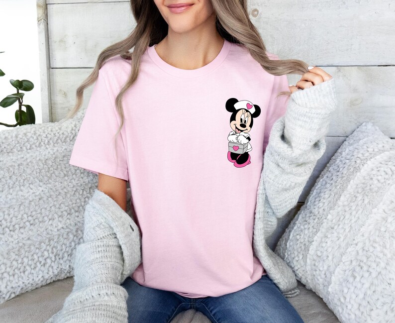 Puede incluir: Una camiseta rosa claro con un dise&ntilde;o de Minnie Mouse. El personaje de dibujos animados lleva un gorro de enfermera blanco con un coraz&oacute;n rosa, sosteniendo una caja gris con un coraz&oacute;n rosa. La camiseta es de color liso.