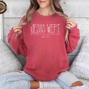 May include: A red-toned sweatshirt with the text "JESUS WEPT" in white, with the words "but He never complained" and "John 11:35" below. The sweatshirt is a solid color and has a crew neck.
