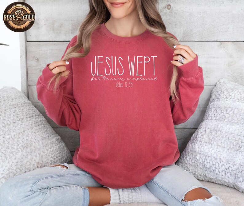 May include: A red-toned sweatshirt with the text "JESUS WEPT" in white, with the words "but He never complained" and "John 11:35" below. The sweatshirt is a solid color and has a crew neck.