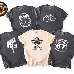 May include: Five t-shirts with various white graphics on them. The graphics include an alarm clock, dice, a street sign, a sombrero with the text "SEIS SIETE", and a Route 67 sign. The shirts are in shades of gray and beige.