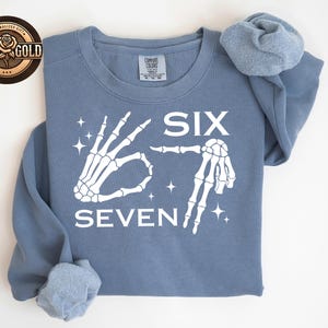 May include: A dusty blue sweatshirt with the words "SIX SEVEN" in white, accompanied by skeleton hand illustrations. The design includes star accents. The sweatshirt has a Comfort Colors tag.