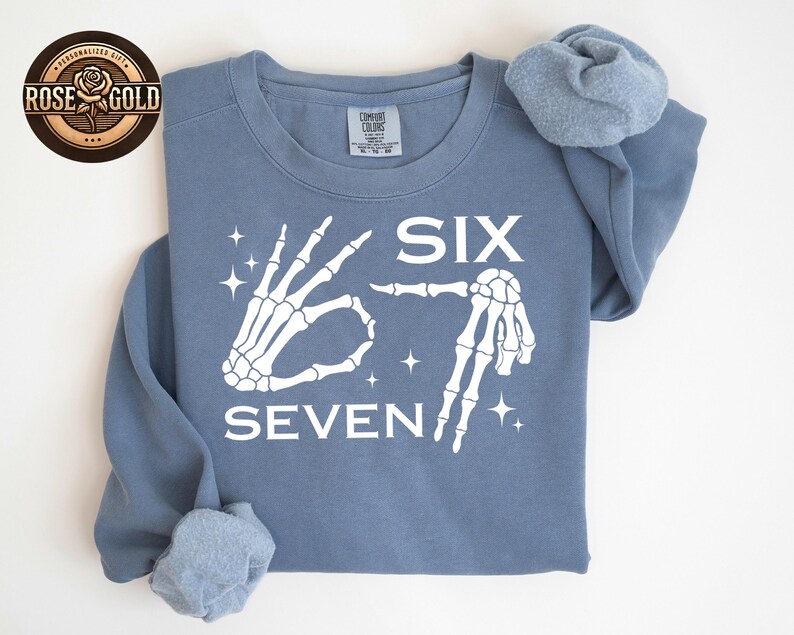 May include: A dusty blue sweatshirt with the words "SIX SEVEN" in white, accompanied by skeleton hand illustrations. The design includes star accents. The sweatshirt has a Comfort Colors tag.