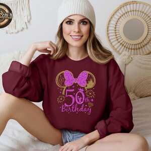 May include: Burgundy sweatshirt with a gold and pink glitter design. The design features a bow, mouse ears, the number "50", and the words "Birthday". The sweatshirt is a long-sleeved, crew neck style.