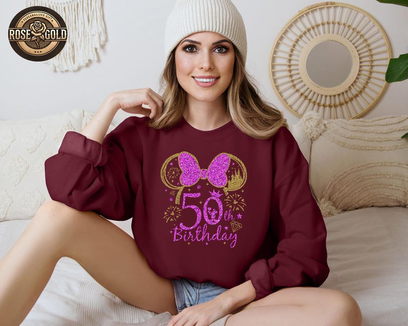 May include: Burgundy sweatshirt with a gold and pink glitter design. The design features a bow, mouse ears, the number "50", and the words "Birthday". The sweatshirt is a long-sleeved, crew neck style.