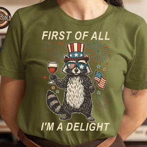 May include: Olive green t-shirt featuring a raccoon wearing sunglasses, a patriotic hat, and holding a wine glass and American flag. The text reads "FIRST OF ALL I'M A DELIGHT".