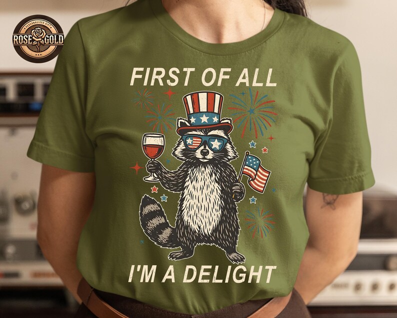 May include: Olive green t-shirt featuring a raccoon wearing sunglasses, a patriotic hat, and holding a wine glass and American flag. The text reads "FIRST OF ALL I'M A DELIGHT".