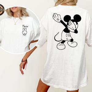 May include: White t-shirt with a black and white Mickey Mouse graphic on the back, in a dabbing pose. The front of the shirt has a peace sign and the name "Taylor". The t-shirt has a relaxed fit.