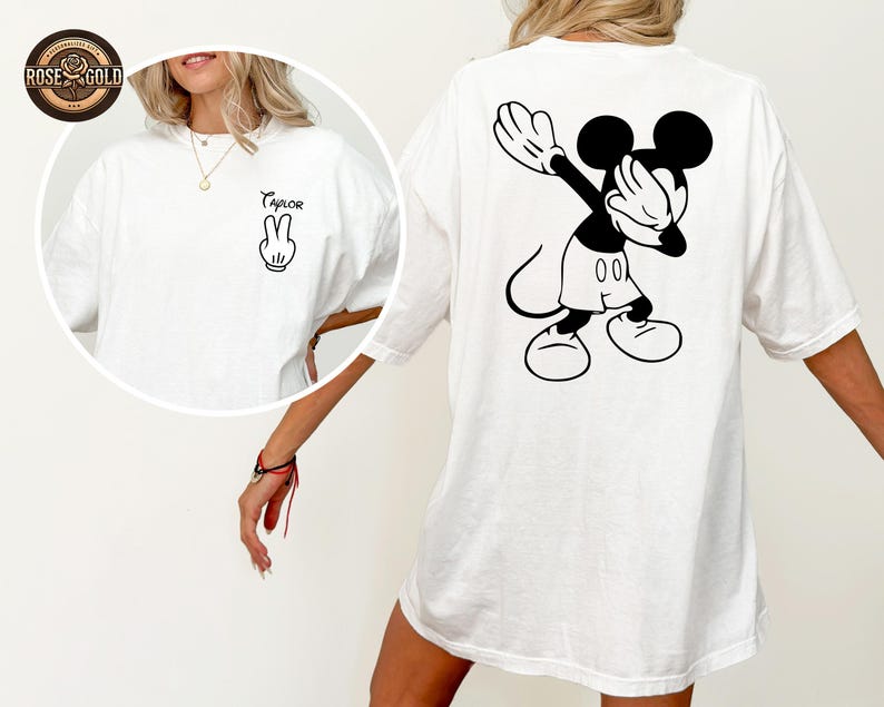 May include: White t-shirt with a black and white Mickey Mouse graphic on the back, in a dabbing pose. The front of the shirt has a peace sign and the name "Taylor". The t-shirt has a relaxed fit.