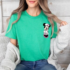 Puede incluir: Camiseta verde con un dise&ntilde;o de Minnie Mouse enfermera sosteniendo una caja con corazones. El dise&ntilde;o utiliza negro, blanco, rosa y rojo. La camiseta es de cuello redondo.