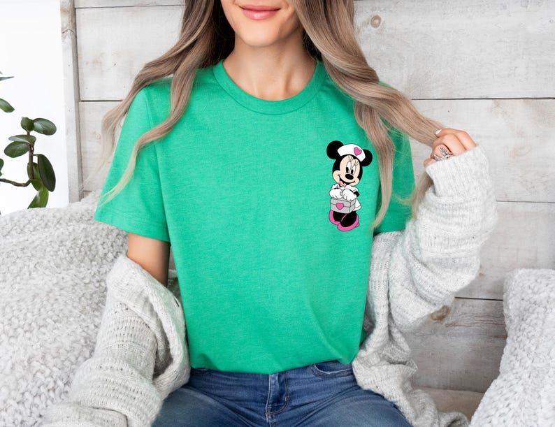 Puede incluir: Camiseta verde con un dise&ntilde;o de Minnie Mouse enfermera sosteniendo una caja con corazones. El dise&ntilde;o utiliza negro, blanco, rosa y rojo. La camiseta es de cuello redondo.