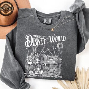 May include: Dark grey sweatshirt featuring a Walt Disney World graphic. The design includes the words "Walt Disney World" and "The Happiest Place on Earth" with illustrations of Mickey Mouse and park attractions. A gold necklace is draped on the sweatshirt.