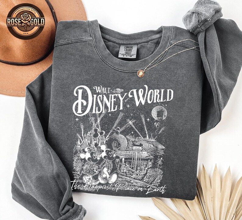 May include: Dark grey sweatshirt featuring a Walt Disney World graphic. The design includes the words "Walt Disney World" and "The Happiest Place on Earth" with illustrations of Mickey Mouse and park attractions. A gold necklace is draped on the sweatshirt.