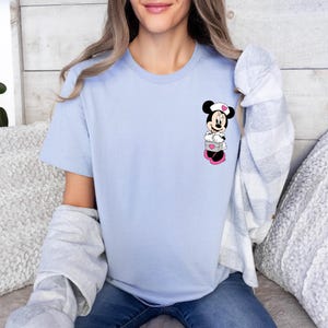 Puede incluir: Camiseta azul claro con un dise&ntilde;o de dibujos animados de Minnie Mouse. El gr&aacute;fico muestra a Minnie Mouse con un gorro de enfermera blanco y zapatos rosas, sosteniendo una caja con corazones. La camiseta es de color azul claro liso.