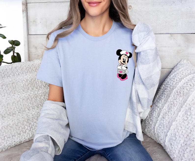 Puede incluir: Camiseta azul claro con un dise&ntilde;o de dibujos animados de Minnie Mouse. El gr&aacute;fico muestra a Minnie Mouse con un gorro de enfermera blanco y zapatos rosas, sosteniendo una caja con corazones. La camiseta es de color azul claro liso.