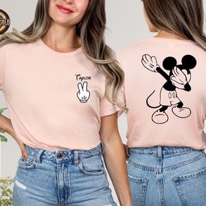 May include: A pale pink t-shirt with a black and white Mickey Mouse graphic on the back. The front displays a peace sign and the name "Taylor". The tee is made of a soft fabric, suitable for everyday wear. The shirt is a casual style.