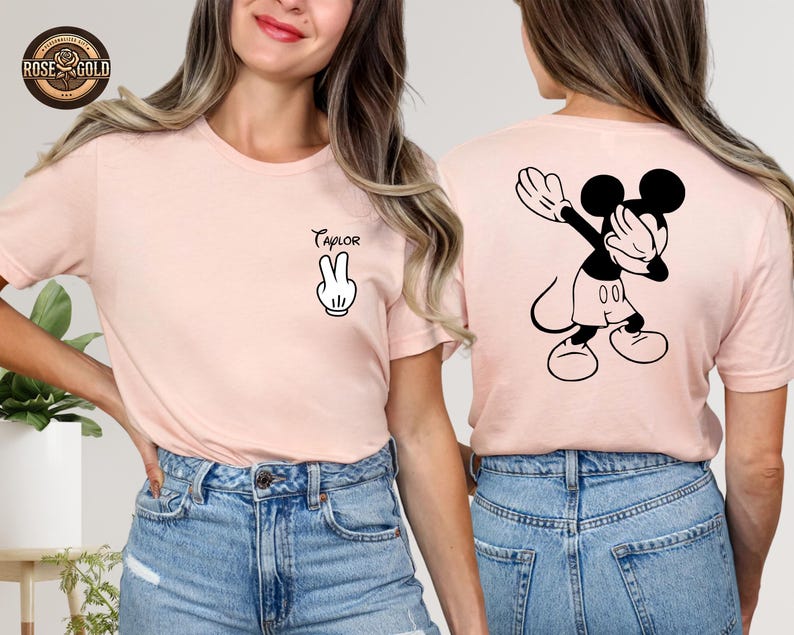 May include: A pale pink t-shirt with a black and white Mickey Mouse graphic on the back. The front displays a peace sign and the name "Taylor". The tee is made of a soft fabric, suitable for everyday wear. The shirt is a casual style.