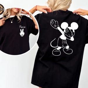 May include: Black t-shirt with a white Mickey Mouse graphic on the back, in a dabbing pose. The front of the shirt has a white peace sign hand and the name "Taylor" in cursive. The shirt has a white collar.